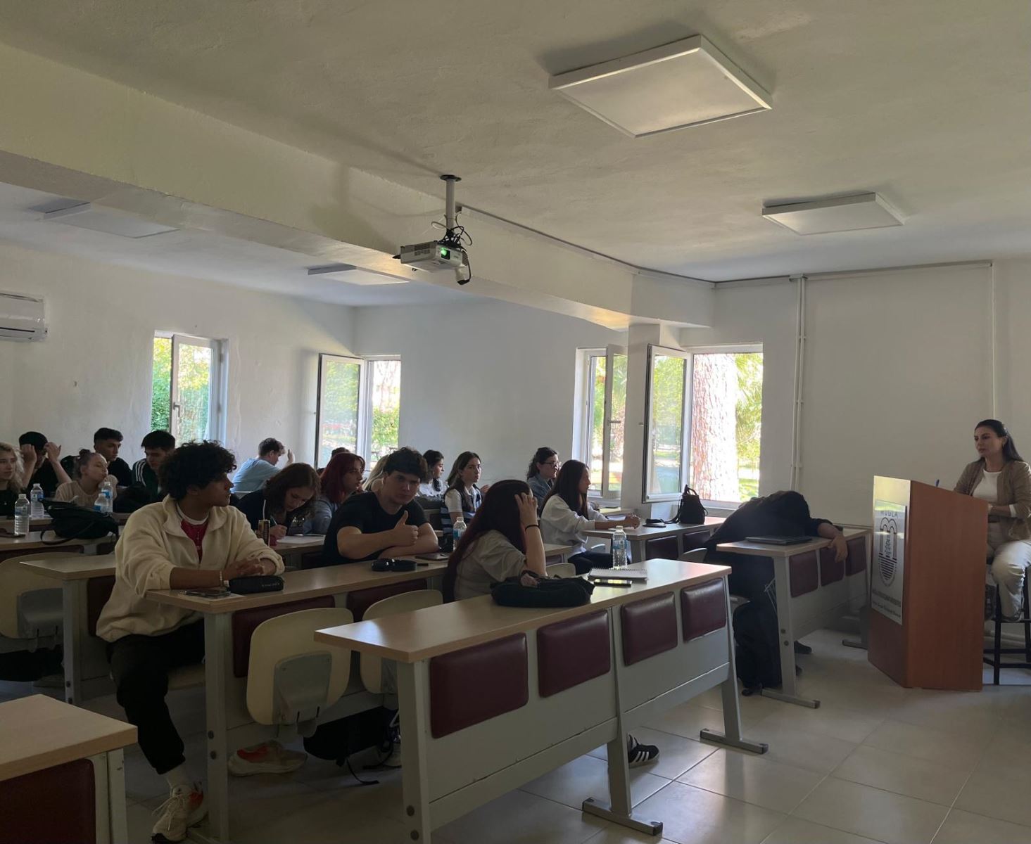 Dalaman Meslek Yüksekokulu sınıfında yapılan toplantı. Ön tarafta kürsüde oturan bir öğretim elemanı konuşma yapıyor. Öğrenciler sıralarda oturmuş, sunumu dinliyor. Pencereler açık; dışarıdan ağaçlık bir alan görünüyor. Aydınlık ve geniş bir sınıf ortamı.